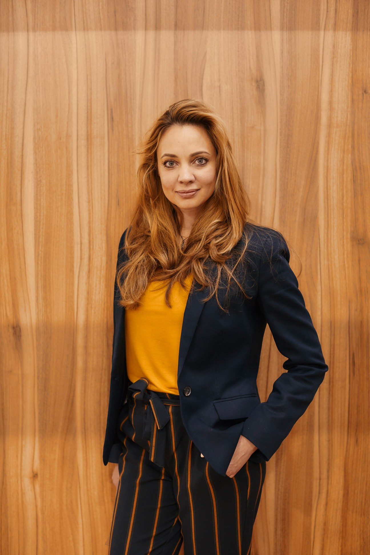 Woman with reddish-brown hair wearing a black blazer and yellow top stands against a wooden backdrop.