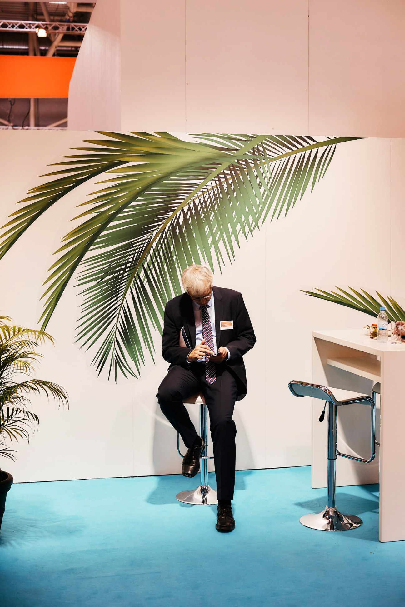 A man in a dark suit sits on a blue stool at an Expo Real booth, with large green palm leaf artwork and tropical plants behind him.