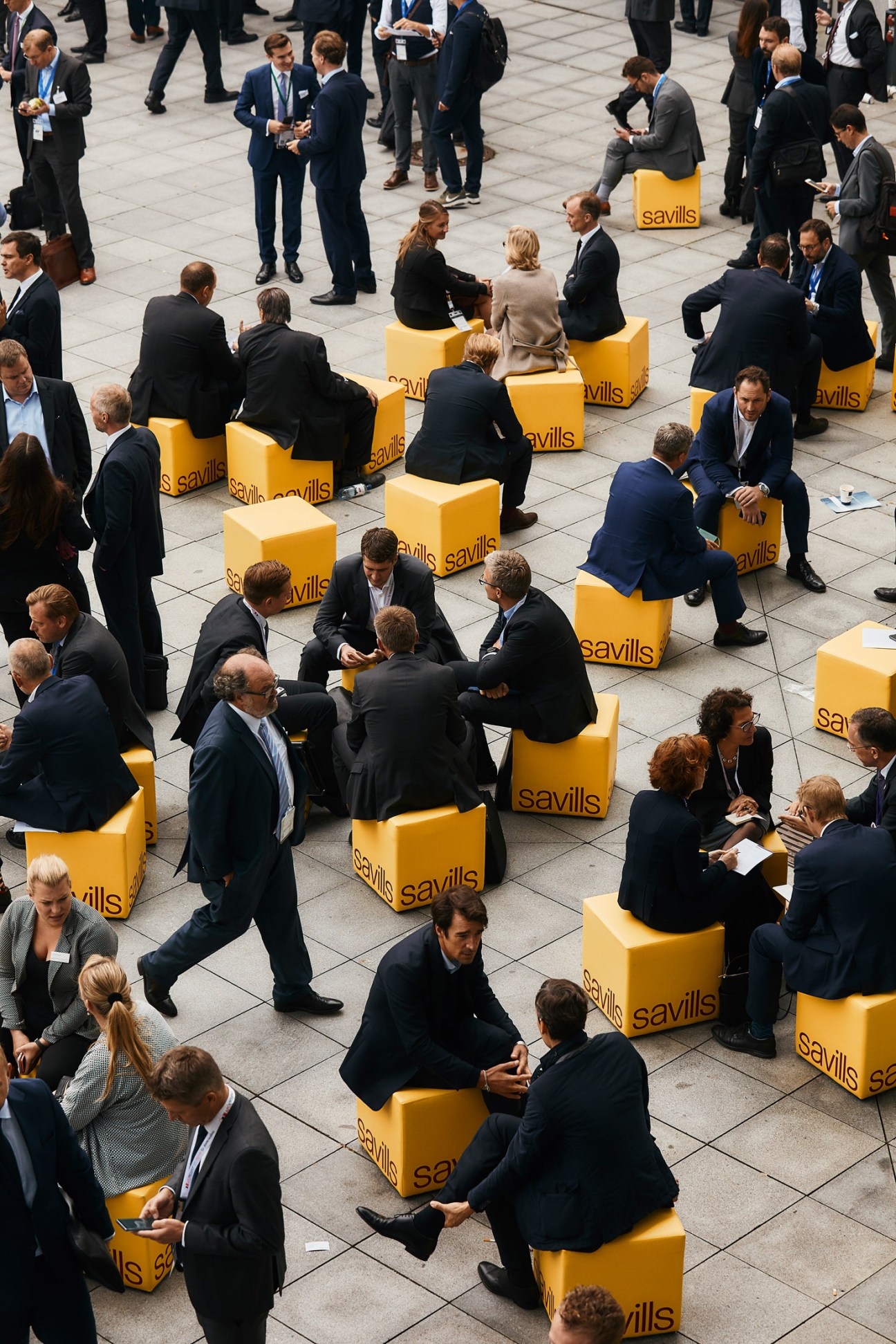 Real estate professionals in business attire conduct informal meetings on yellow branded Savills seats in an outdoor courtyard at Expo Real.