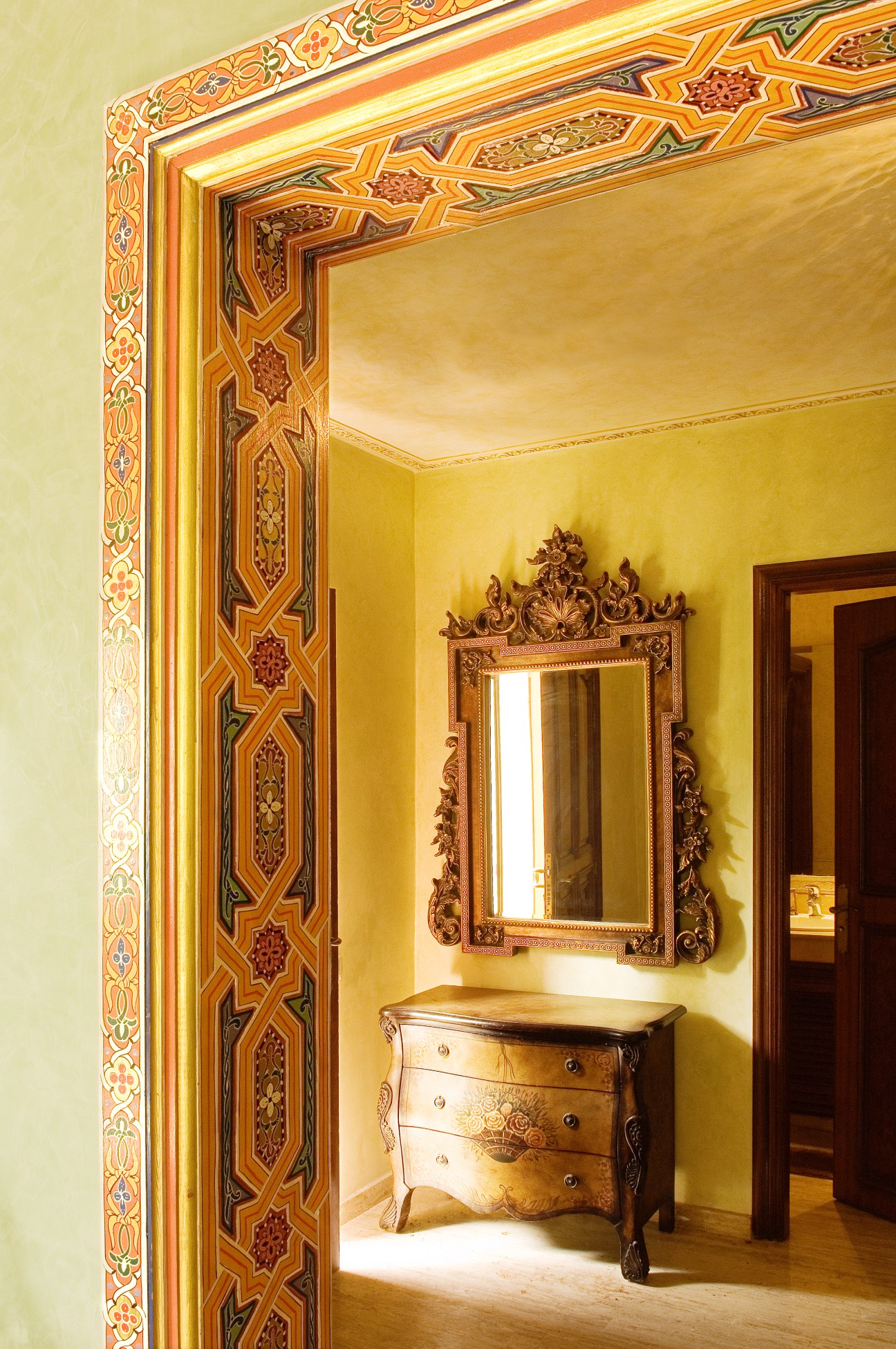 Yellow-walled room with ornate Moroccan geometric wooden doorframe, gilt mirror and decorated wooden dresser.