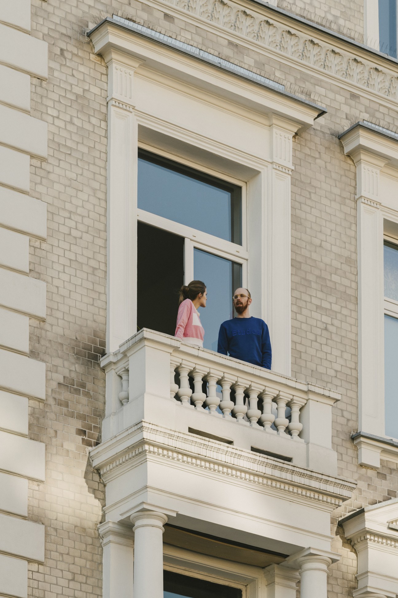 Two people stand on a balcony of a prewar residential building with ornate white trim and brick facade.