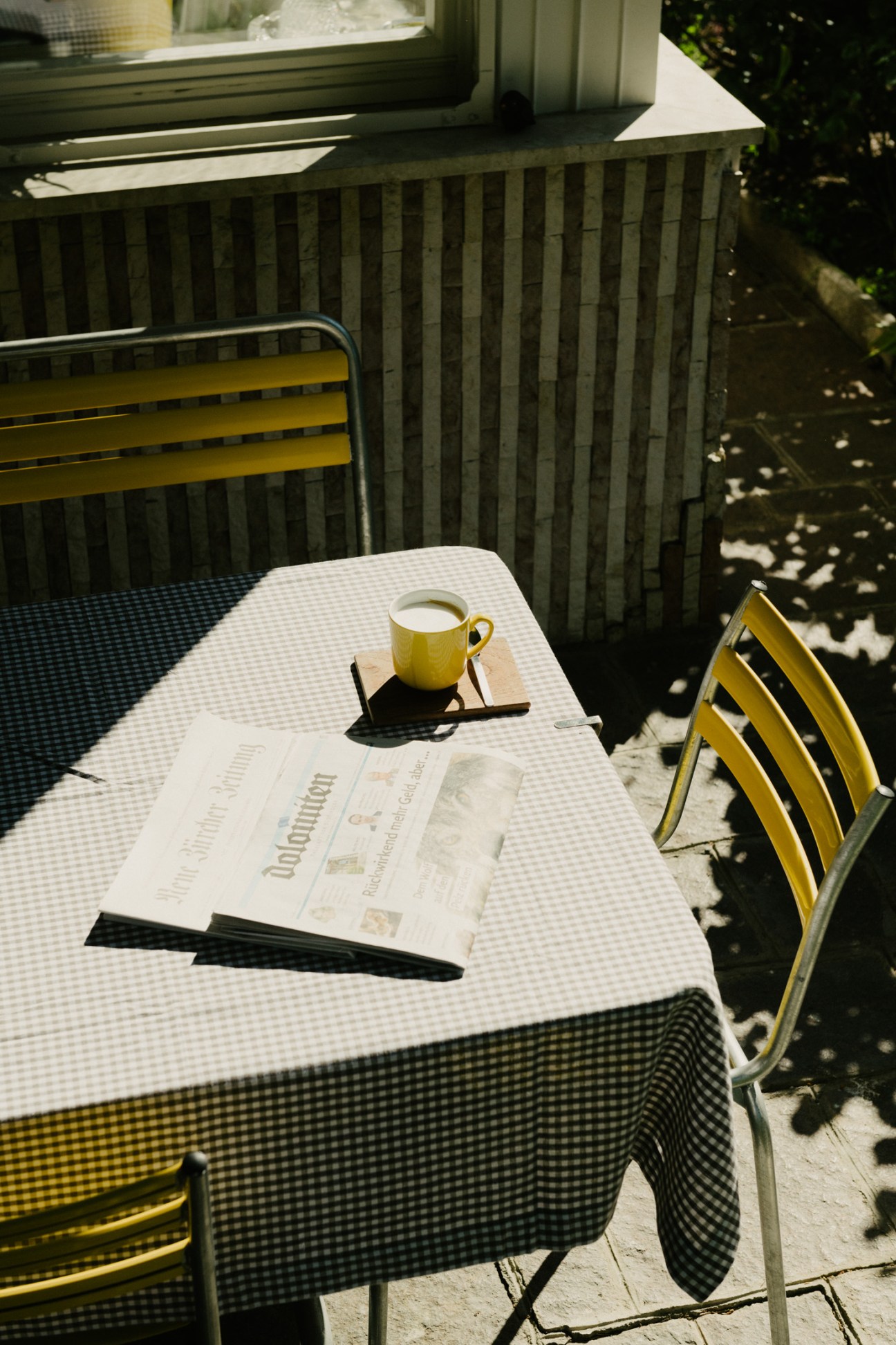 A yellow mug of coffee and newspaper on a checkered table with yellow Manufakt café chairs in sunlight.