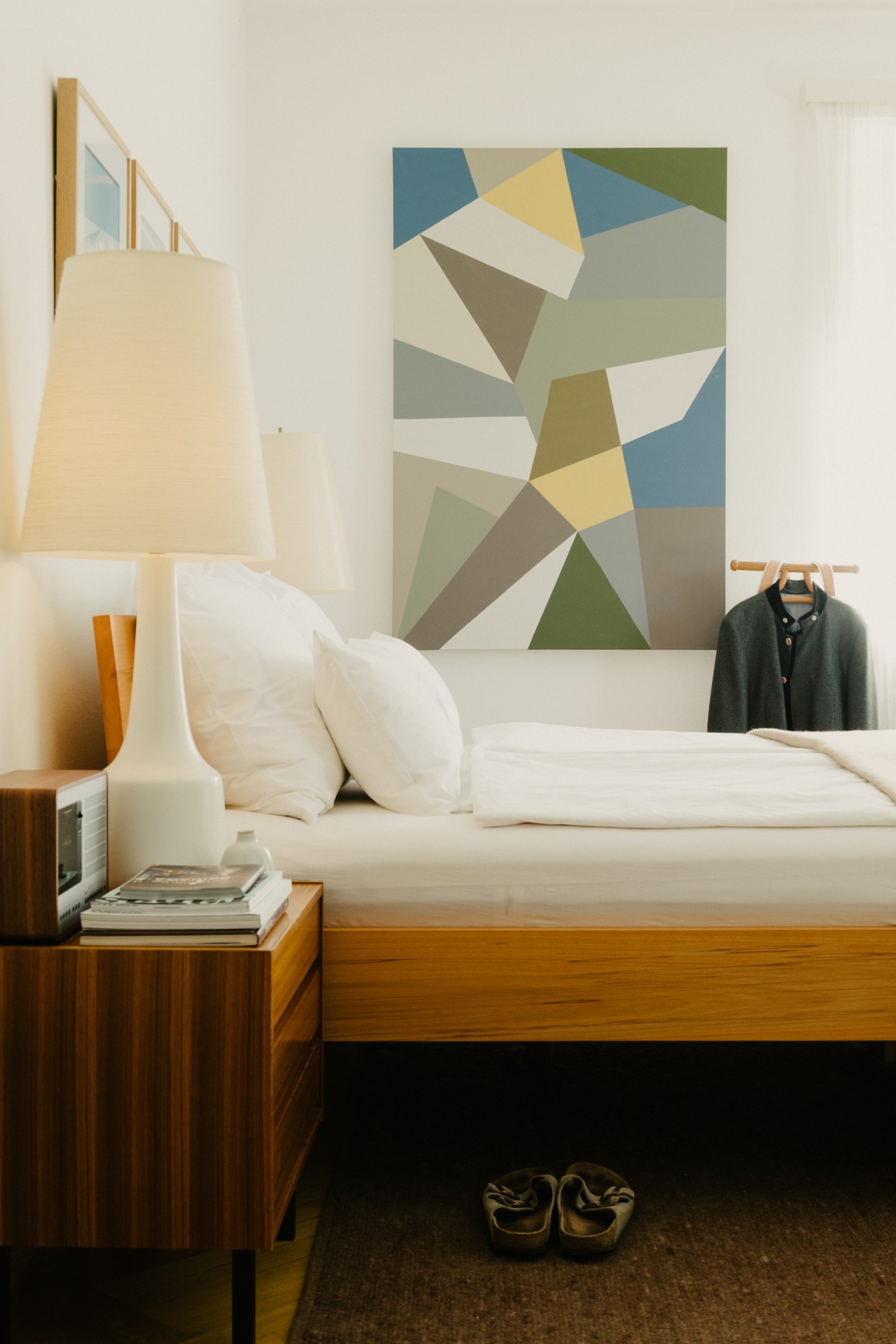 Master bedroom with white bed, wooden nightstand, cream table lamp, and geometric chevron artwork by Virge above headboard.