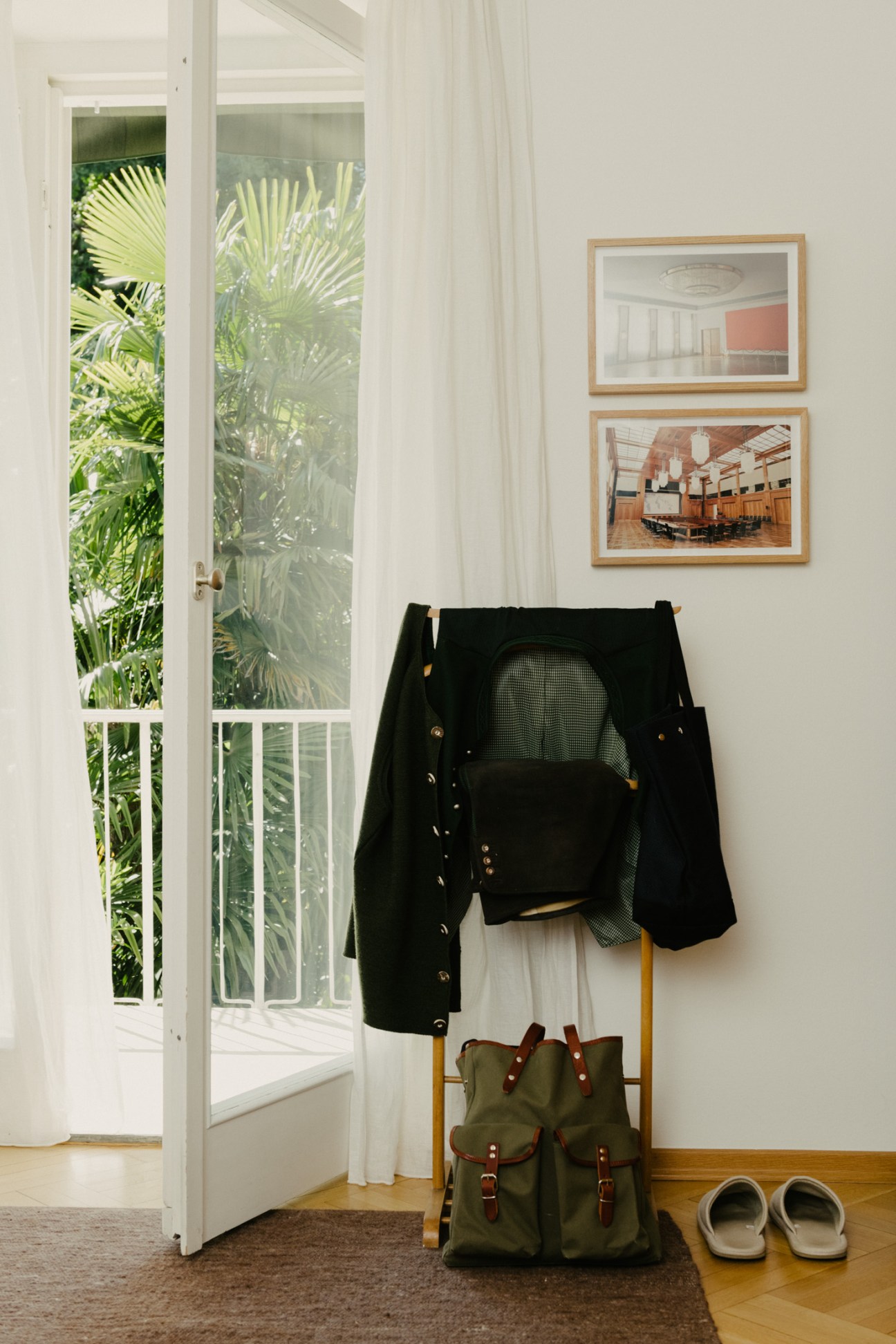Entrance hallway with framed architectural photography prints, a black blazer on a wooden chair, olive canvas rucksack, and white slippers.