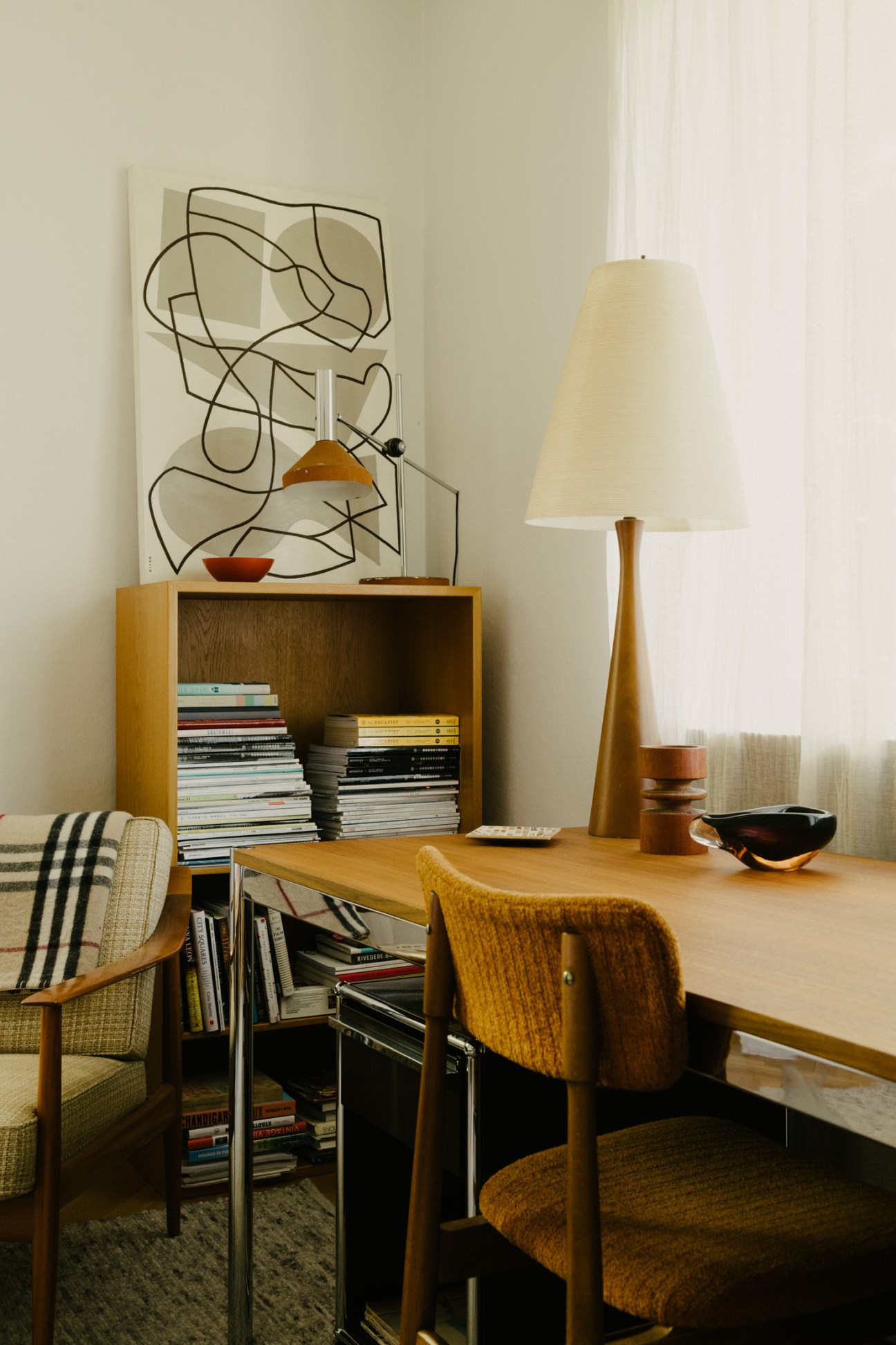 Mid-century modern workspace with wooden desk, upholstered chairs, USM shelving unit filled with books, abstract wall art, and cream table lamp.
