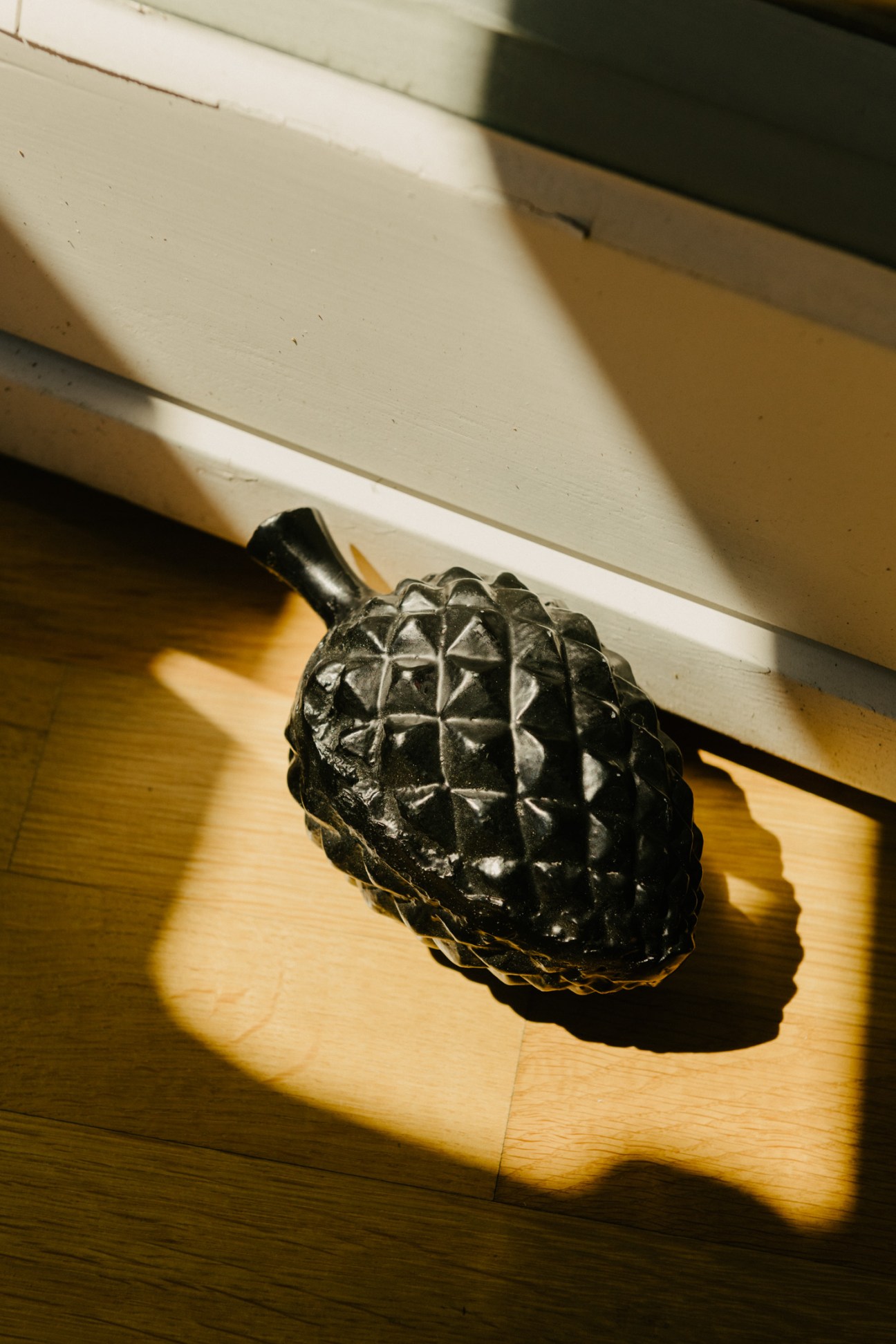 Black metal pine-cone shaped doorstop on wooden floor with white baseboard and sunlight shadow.