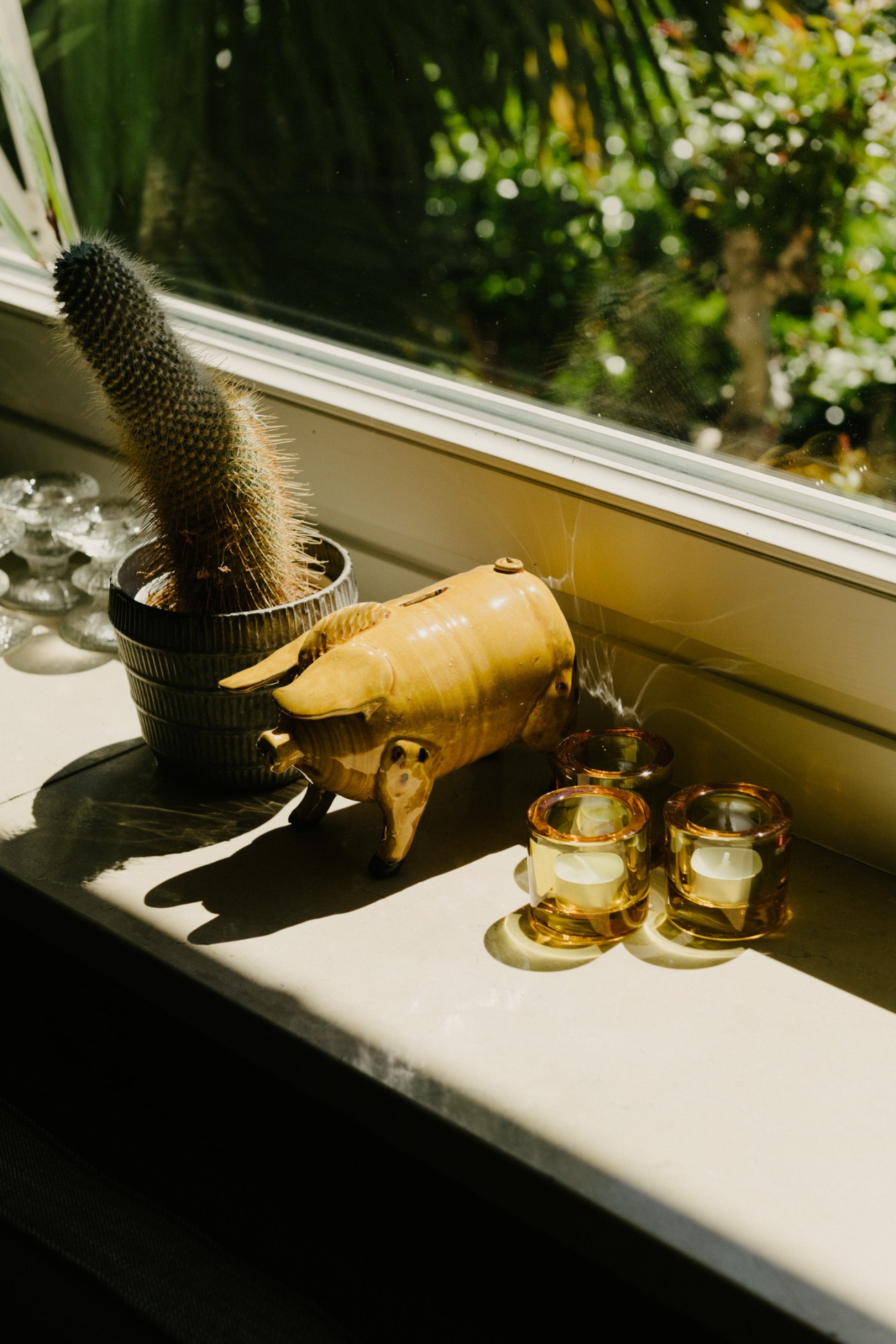 A wooden pig figurine, potted cactus, and amber glass candles displayed on a sunny windowsill.