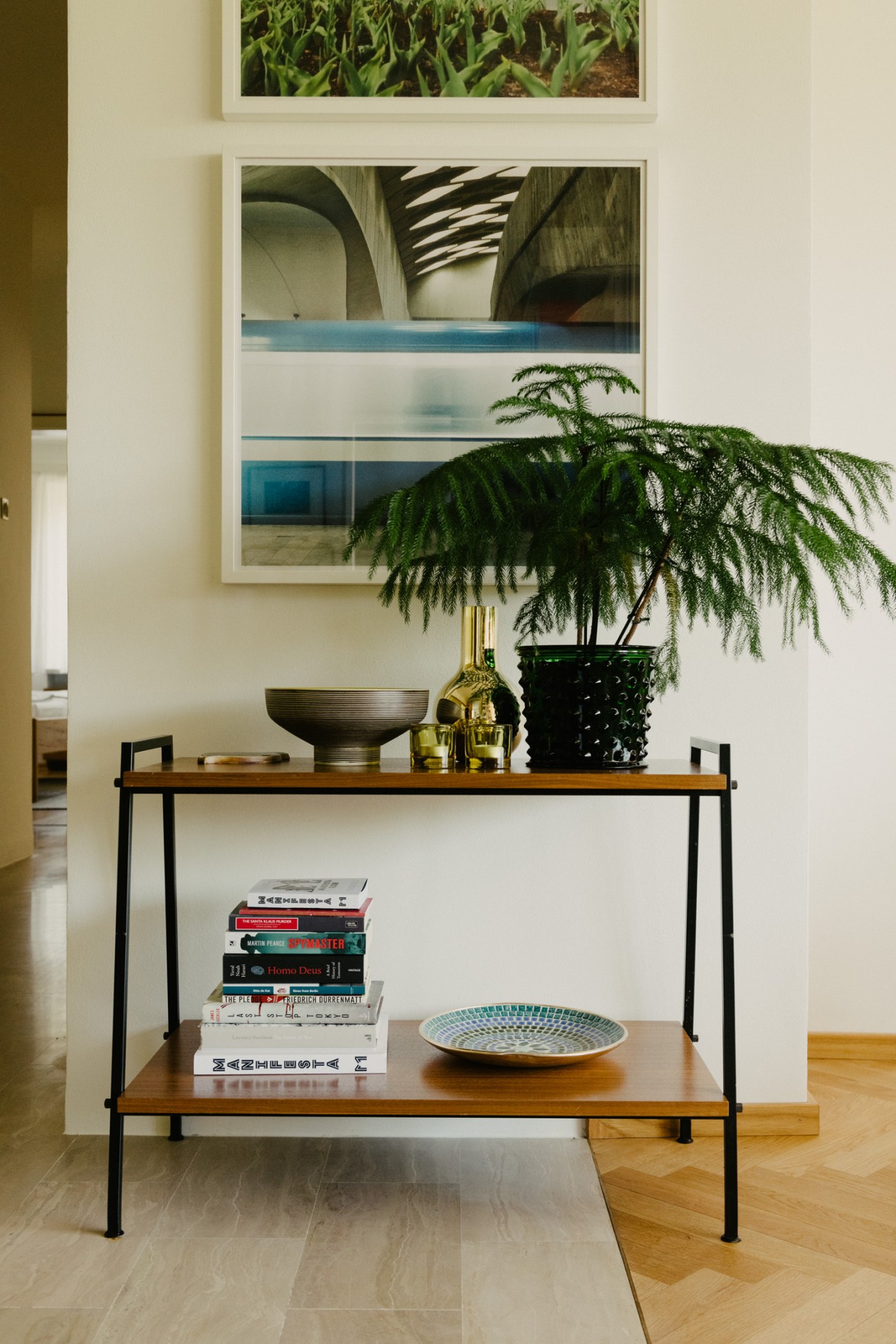 A modernist console table with stacked design books, a ceramic plate, potted fern plant, and framed architectural photograph on white wall.