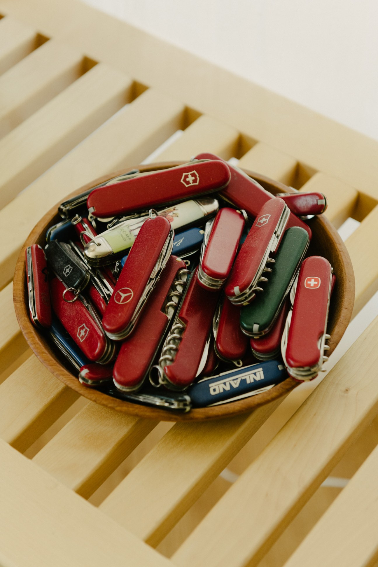 Collection of red and green Swiss Army knives arranged in a wooden bowl on a light wooden shelf.