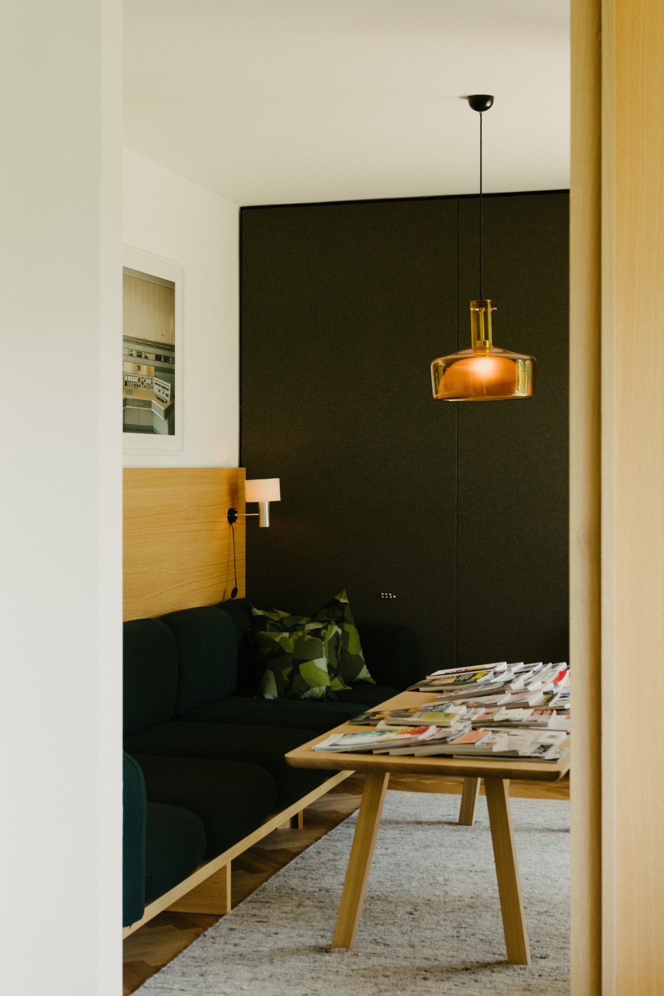 Modern living room with built-in oak couch, black wall panel, wooden coffee table with magazines, and vintage amber glass pendant lamp.