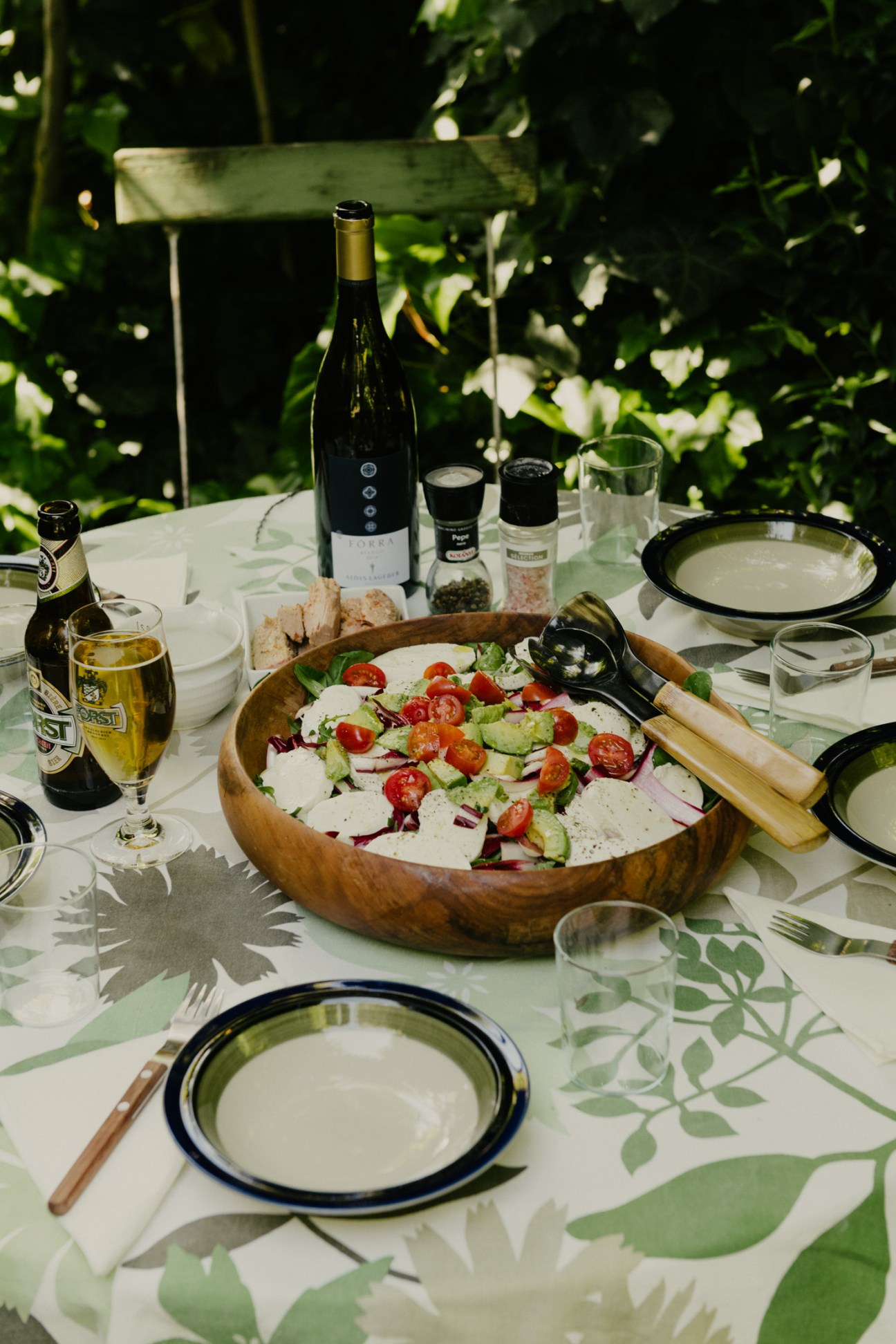 Outdoor dining table with Merano salad in wooden bowl, wine bottle, and Forst beer under vine-covered pergola.