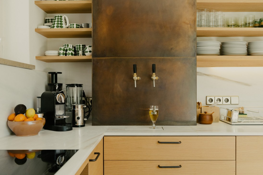 Modern kitchen with bronze metal backsplash, wooden shelving, and a brass beer tap designed by Harry Thaler.