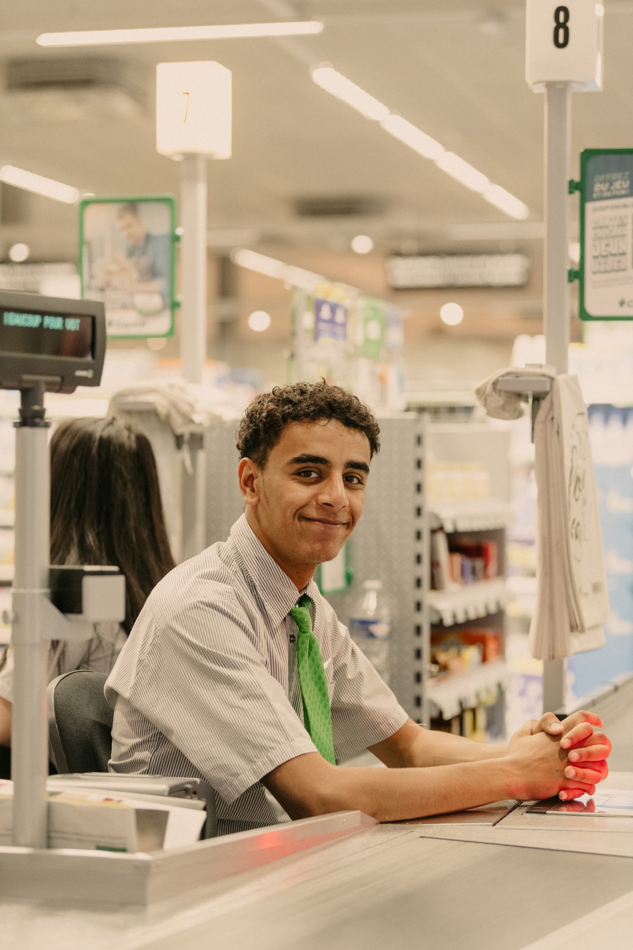 Smiling Cactus supermarket cashier in white shirt and green tie at checkout counter.