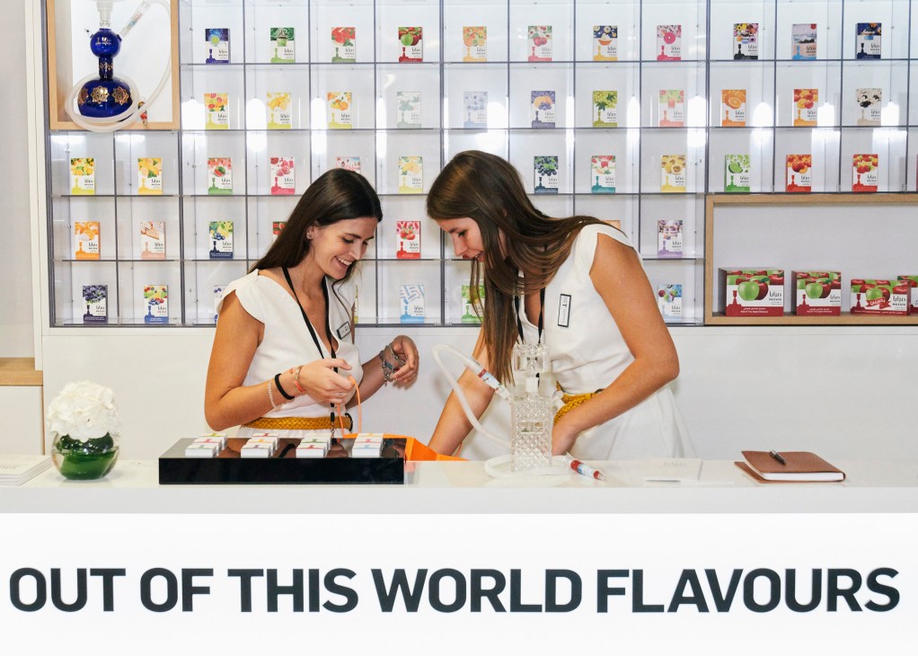 Two women in white examining tea samples at a display counter with colorful tea packages on shelves behind them