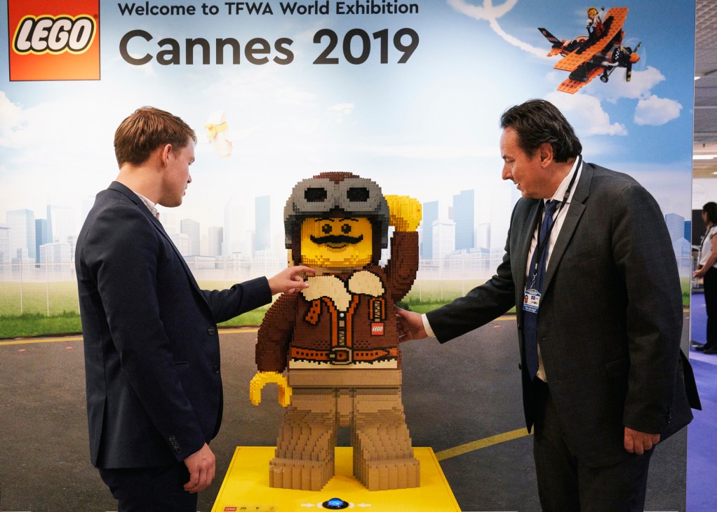 Two businessmen in suits examining a large LEGO minifigure display at TFWA World Exhibition Cannes 2019
