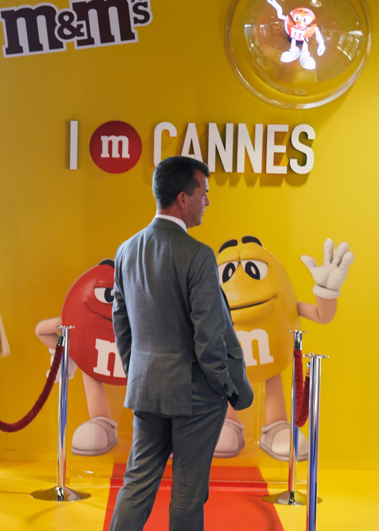 Man in grey suit walking on red carpet past yellow M&M's branded backdrop with cartoon characters