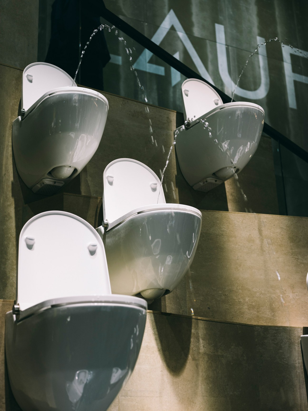 Three modern white toilet seats with water spraying from integrated bidet features mounted on a metallic display wall.
