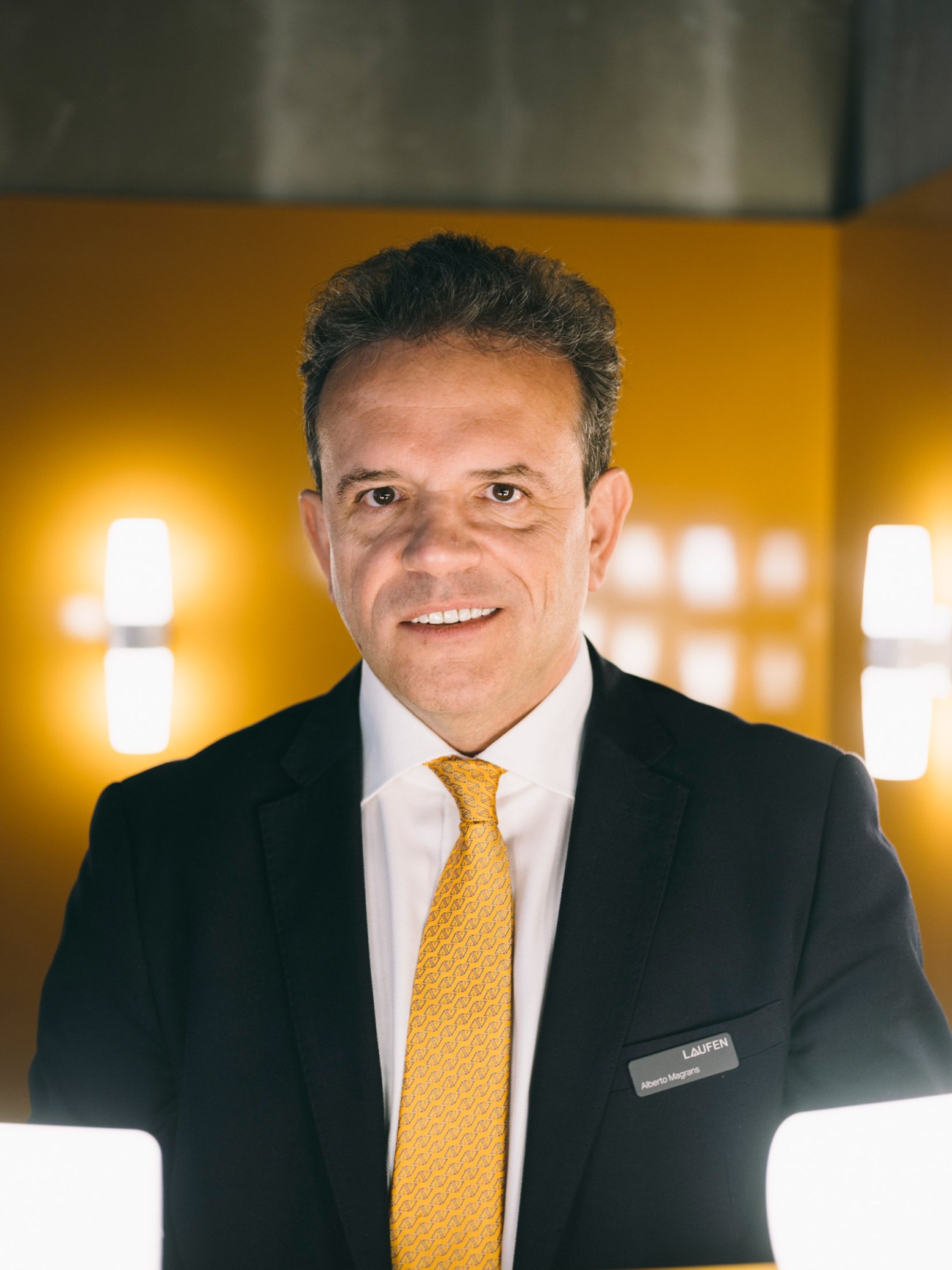 Alberto Magrans, senior managing director of Laufen, wearing a dark suit and gold tie against a yellow backdrop.