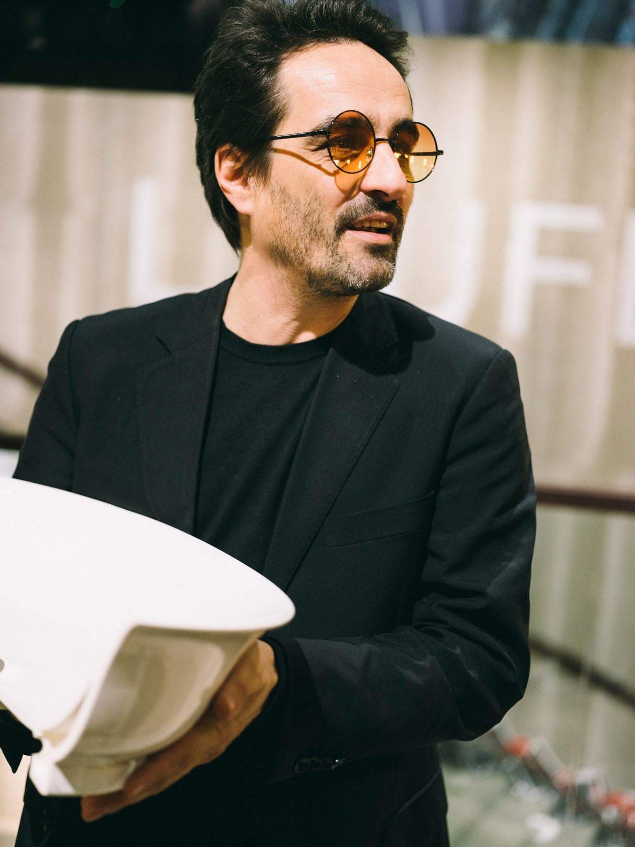 Man wearing round amber-tinted glasses and black blazer holding papers at bathroom design fair.