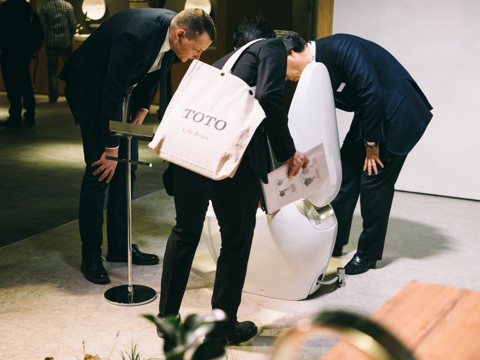 Two men in business suits examine a white TOTO toilet fixture at a trade show display.