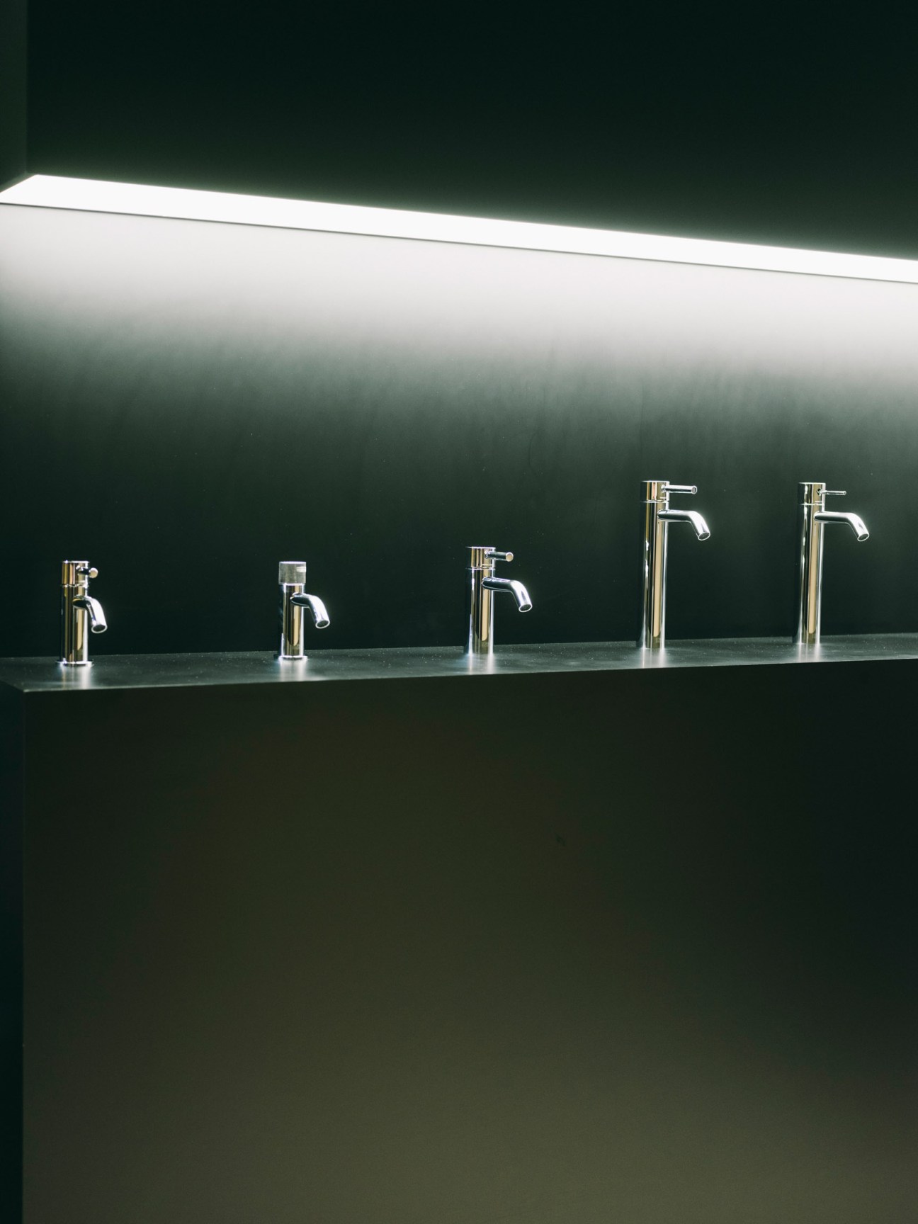 Five chrome taps of varying heights displayed on a counter against a dark green wall with overhead lighting.