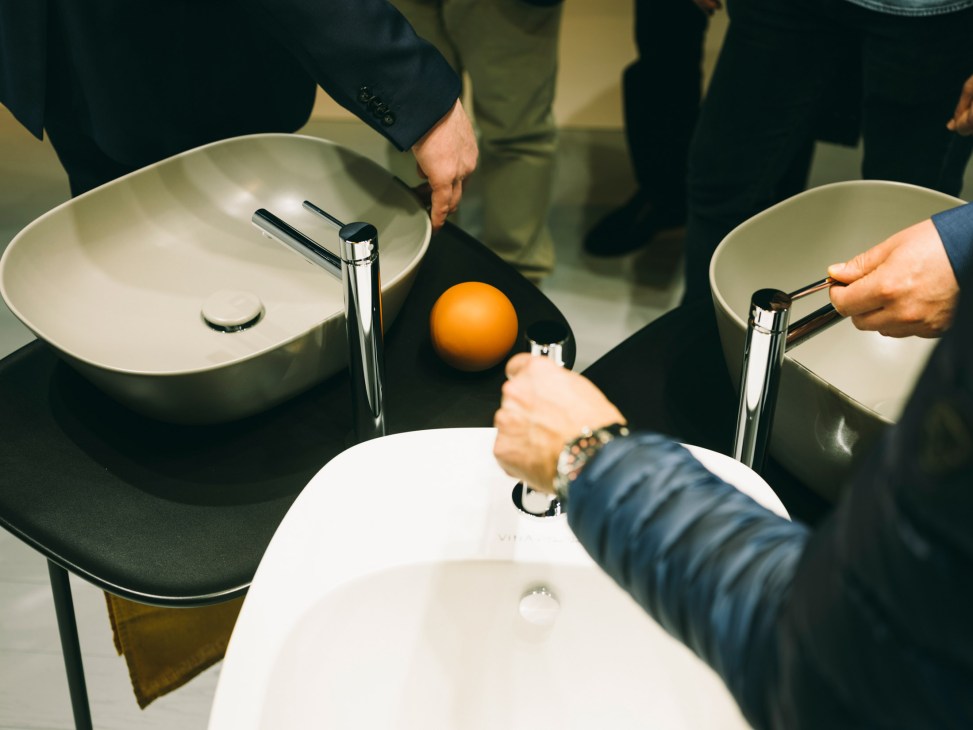 People in business attire holding white ceramic bowls with chrome faucets and an orange sphere displayed between them.