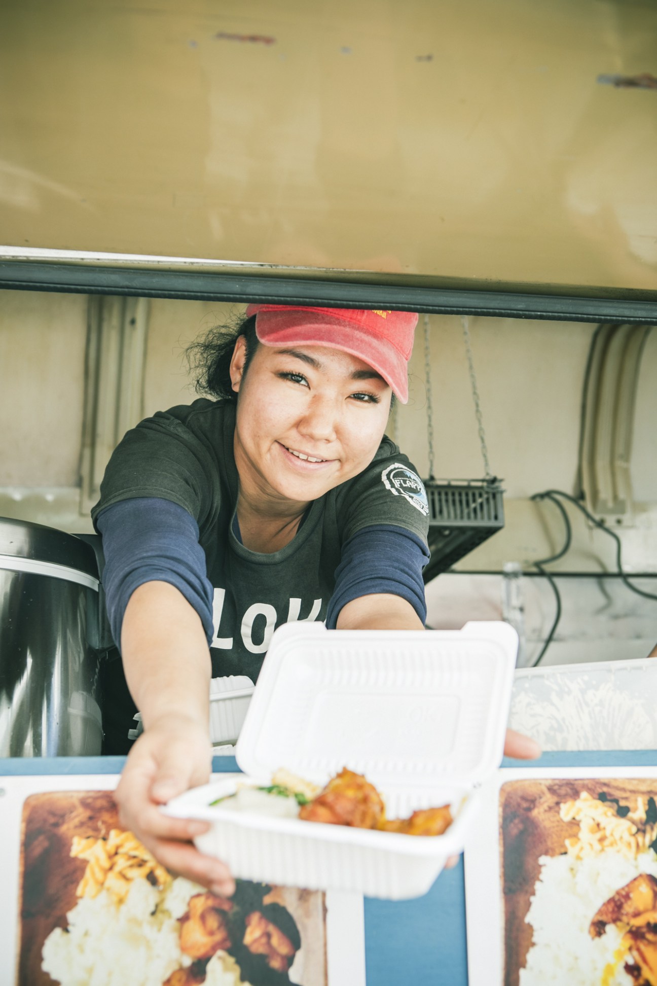 Food truck vendor in red cap smiling while serving Hawaiian-style fried chicken from service window.