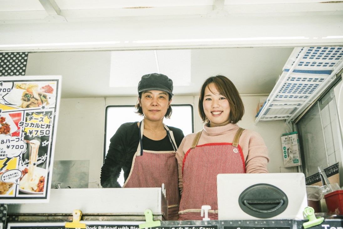 Two women in aprons stand at a food truck service window with menu boards visible above.