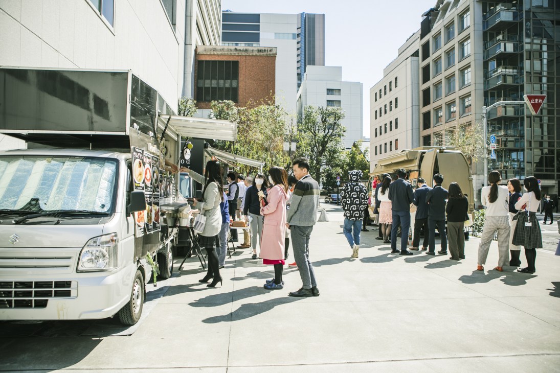 Office workers queue at food trucks parked in a Tokyo street lined with modern buildings.