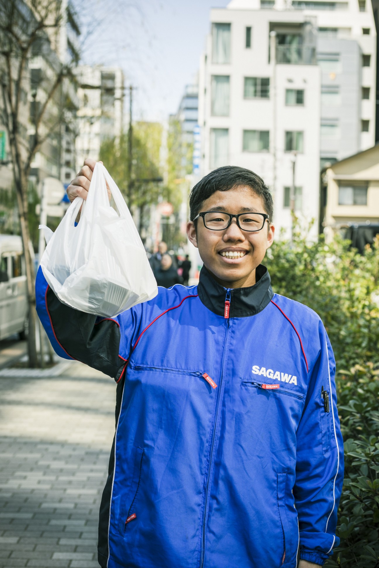 A smiling Sagawa delivery man in blue uniform holds a food package outside Tokyo office buildings.