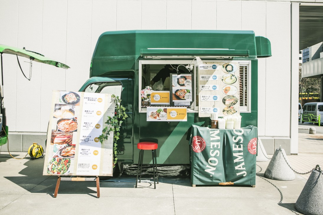 A green food truck with serving window displaying menu boards and offering Italian cuisine from Shodai Kawabe's kitchen car.