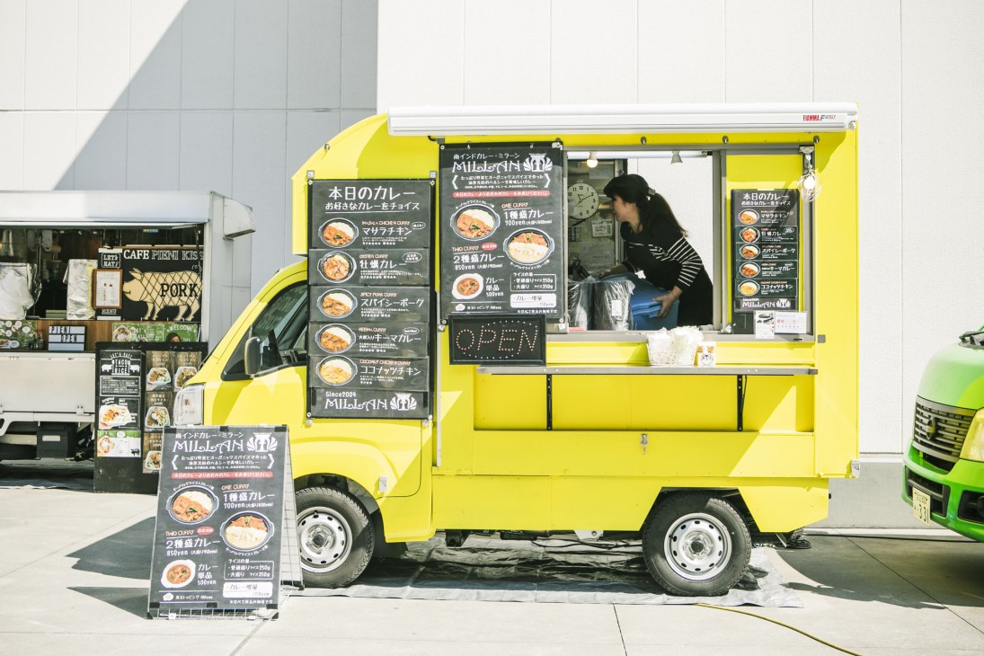 A bright yellow food truck with Japanese signage and menu boards displays curry dishes while a vendor operates from the service window.