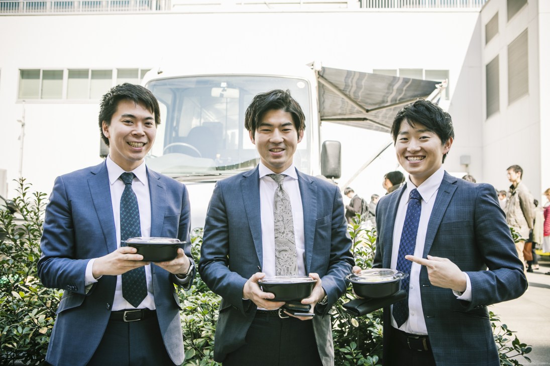 Three smiling men in business suits holding bowls of food in front of a food truck in Tokyo.
