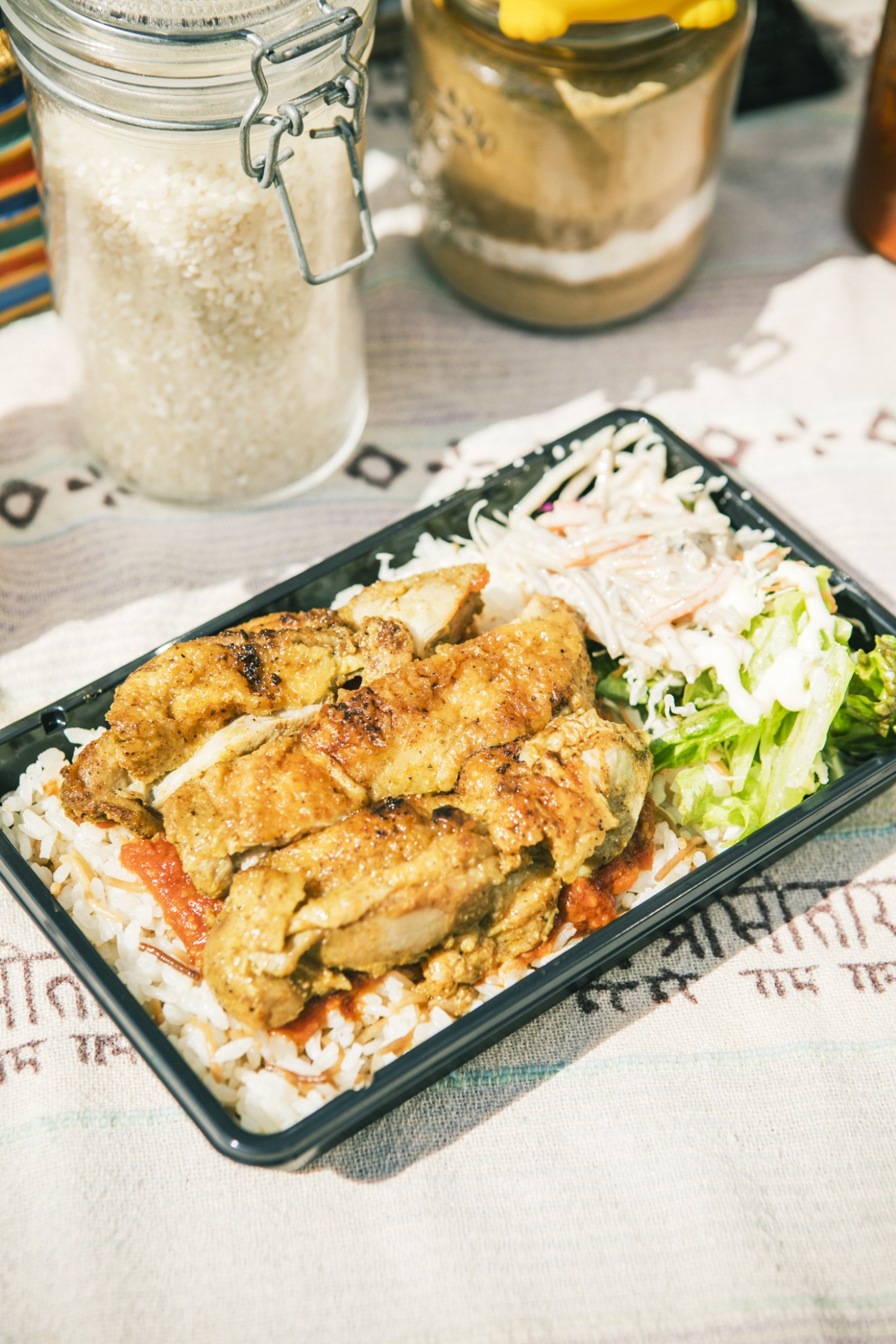 A black bento box containing Egyptian koshary with seasoned chicken, rice, noodles and vegetables, served with glass jars of condiments.
