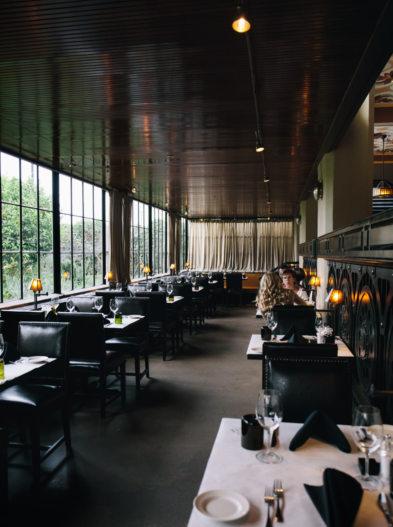 Modern restaurant dining room with black leather chairs, white tablecloths, and large windows overlooking greenery.