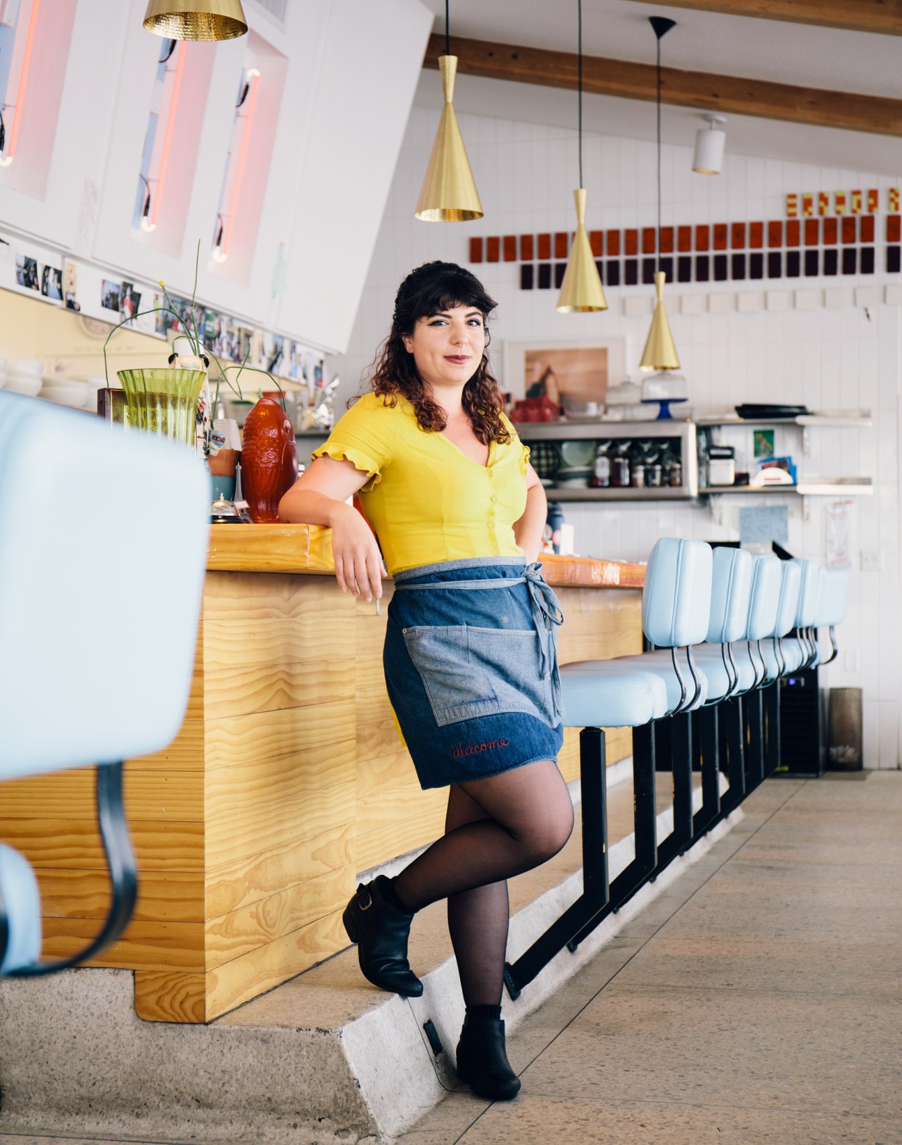 Karen Renda, server at Welcome Diner, poses at the counter of the modern café with brass pendant lights and color swatches on the wall.