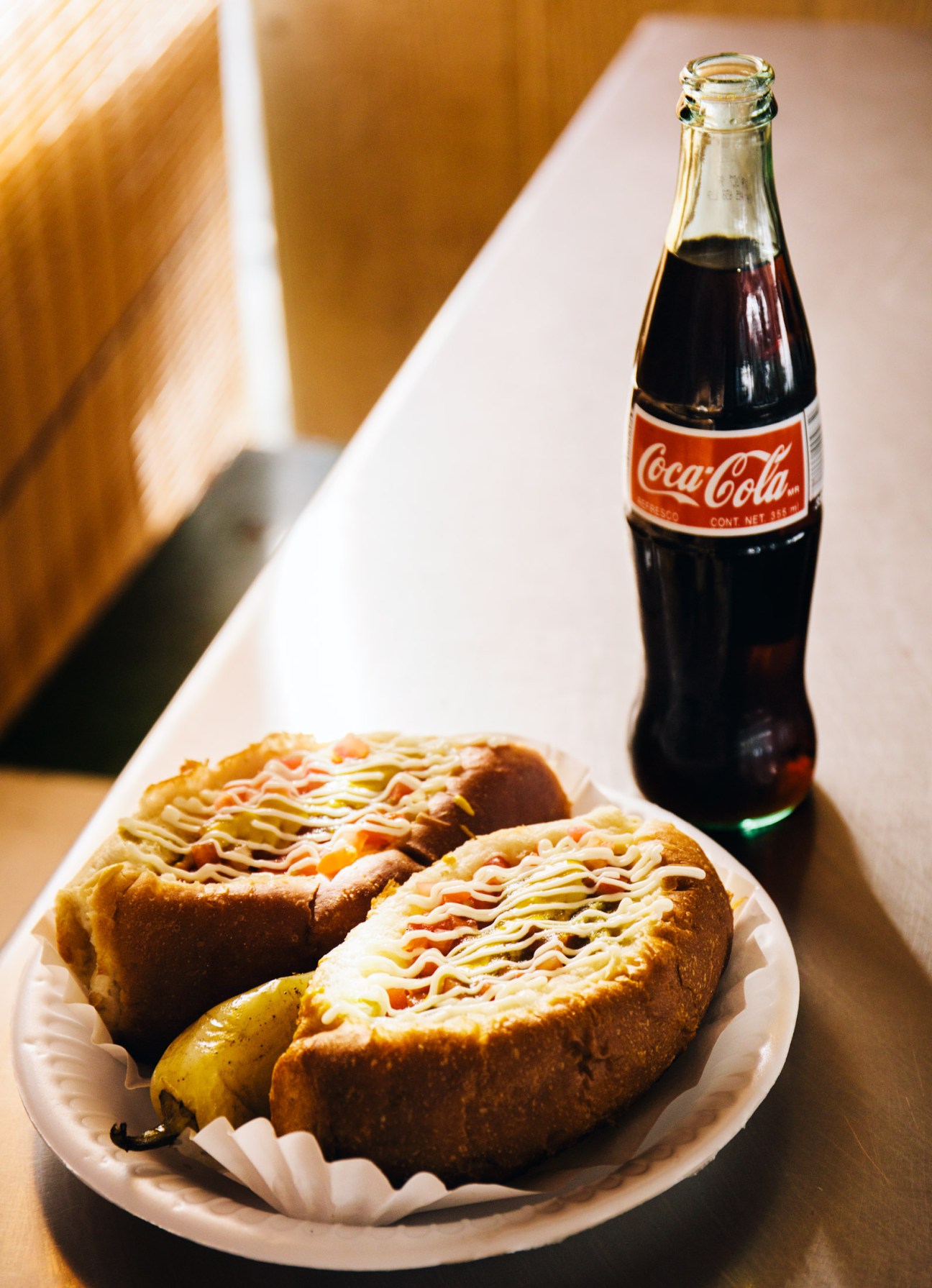 Plate of Sonoran hot dogs with bacon and toppings next to a Coca-Cola bottle.