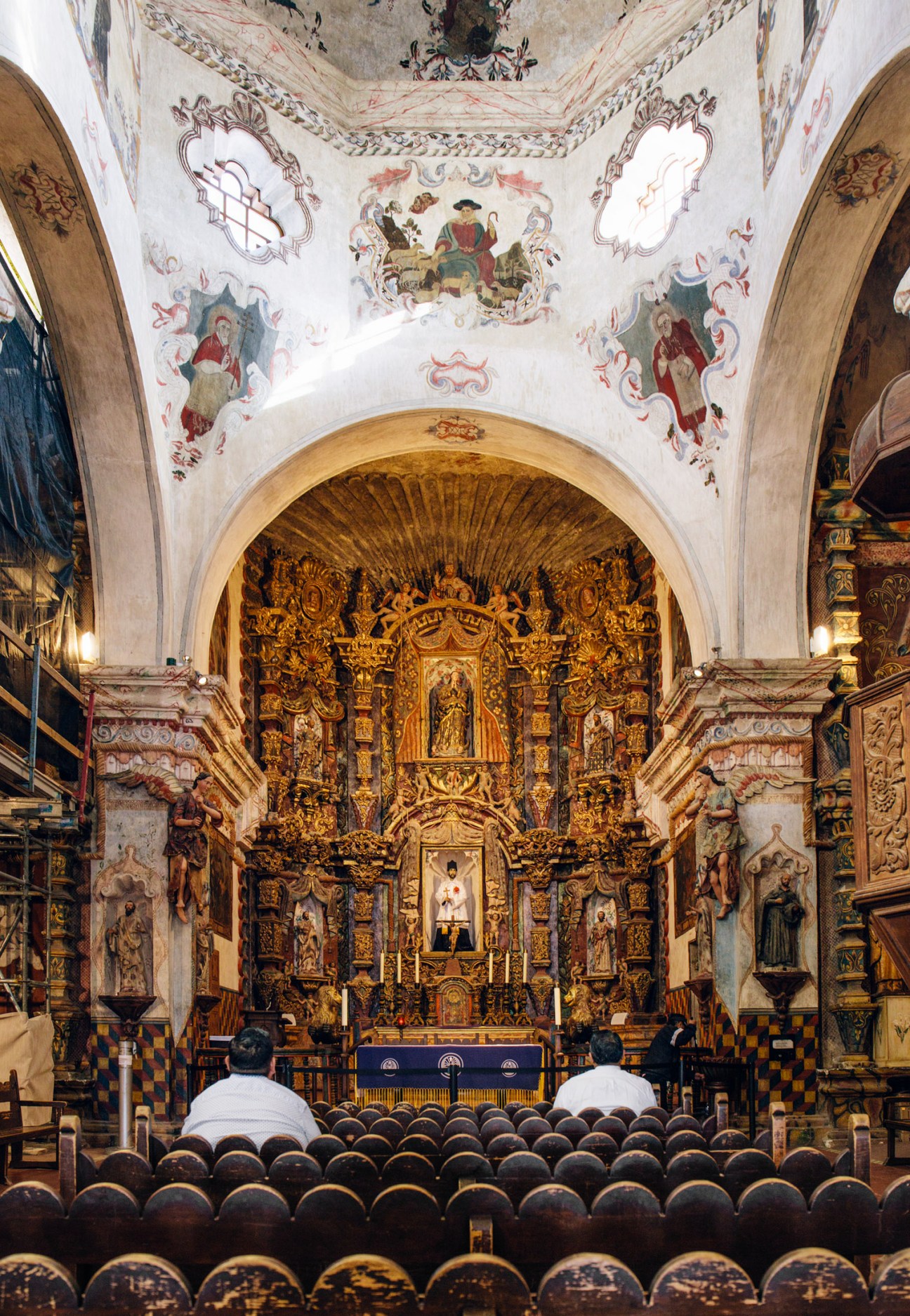 Interior of Mission San Xavier del Bac showing ornate golden altar, white ceiling with religious frescoes, and wooden pews.