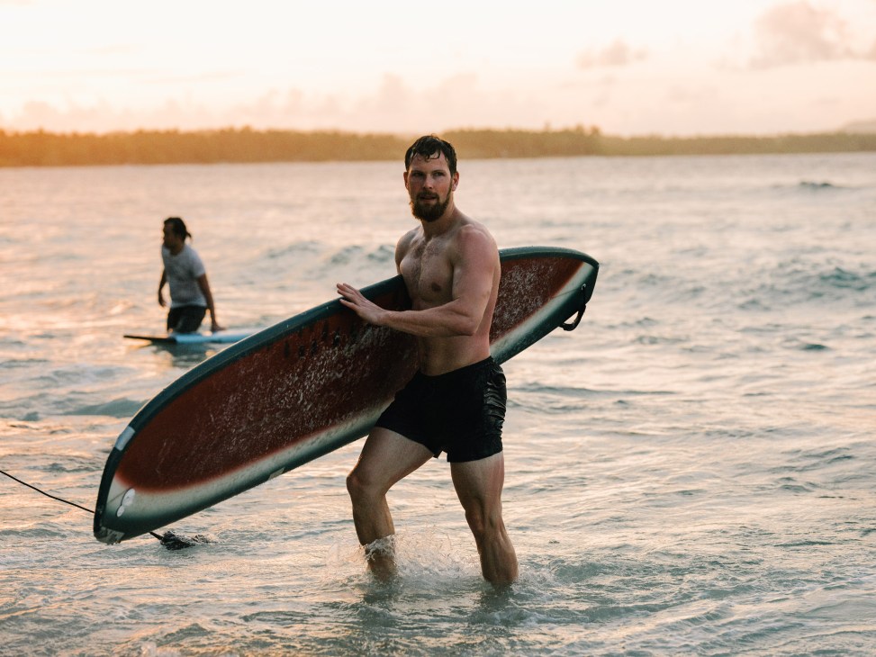 A bearded man carries a surfboard while wading in the ocean at sunset in Siargao.