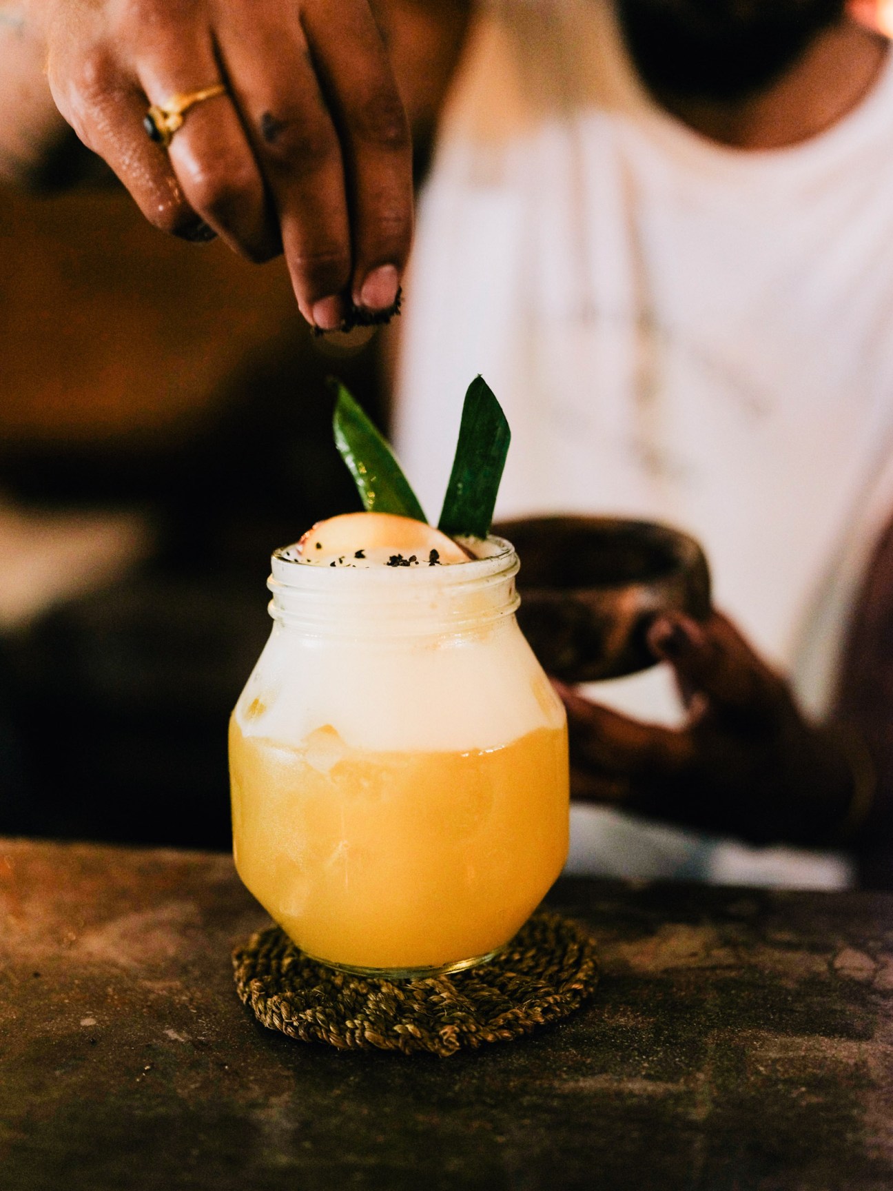 A person's hand garnishes a yellow cocktail with black sesame seeds and green leaves in a glass jar.