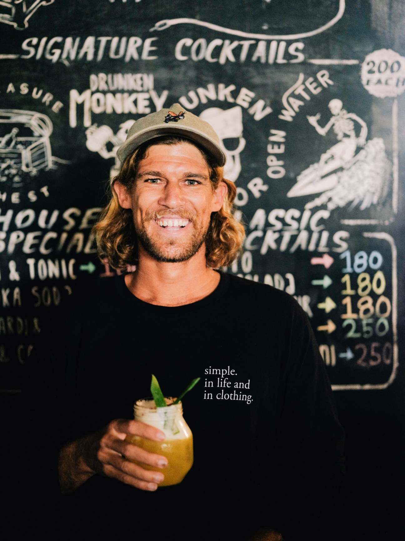 Man with long hair and beard wearing a cap holds a tropical cocktail in front of a signature cocktails menu board.