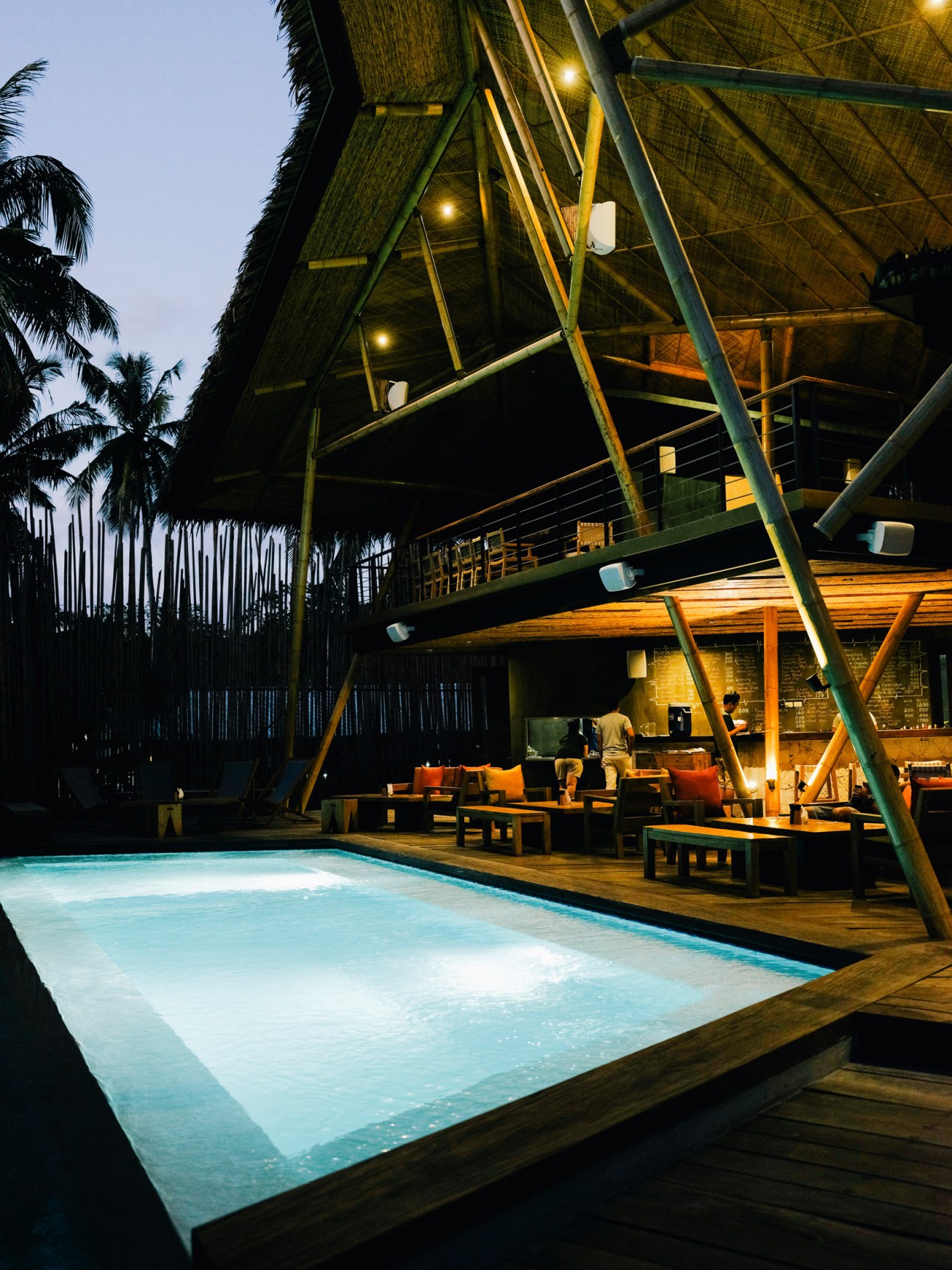 Nighttime view of La Mari bar with illuminated thatch roof, wooden decking, glowing pool, and tropical palm trees.