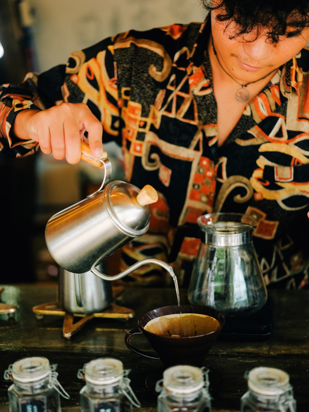 A woman in a patterned orange and black shirt pours hot water from a metal gooseneck kettle into a black coffee cup.