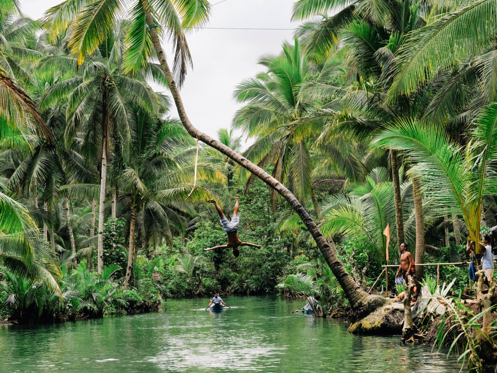 A person swings on a rope above a river surrounded by dense palm trees and tropical vegetation in Siargao, Philippines.
