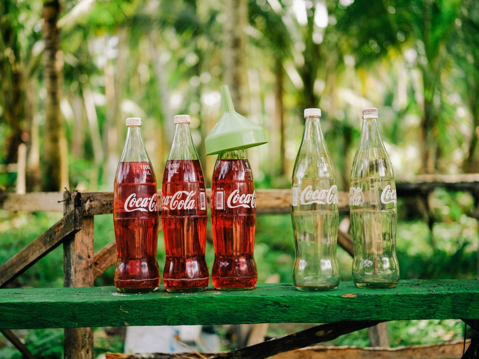 Coca-Cola glass bottles filled with red and clear liquid arranged on a green wooden surface with tropical foliage in the background.