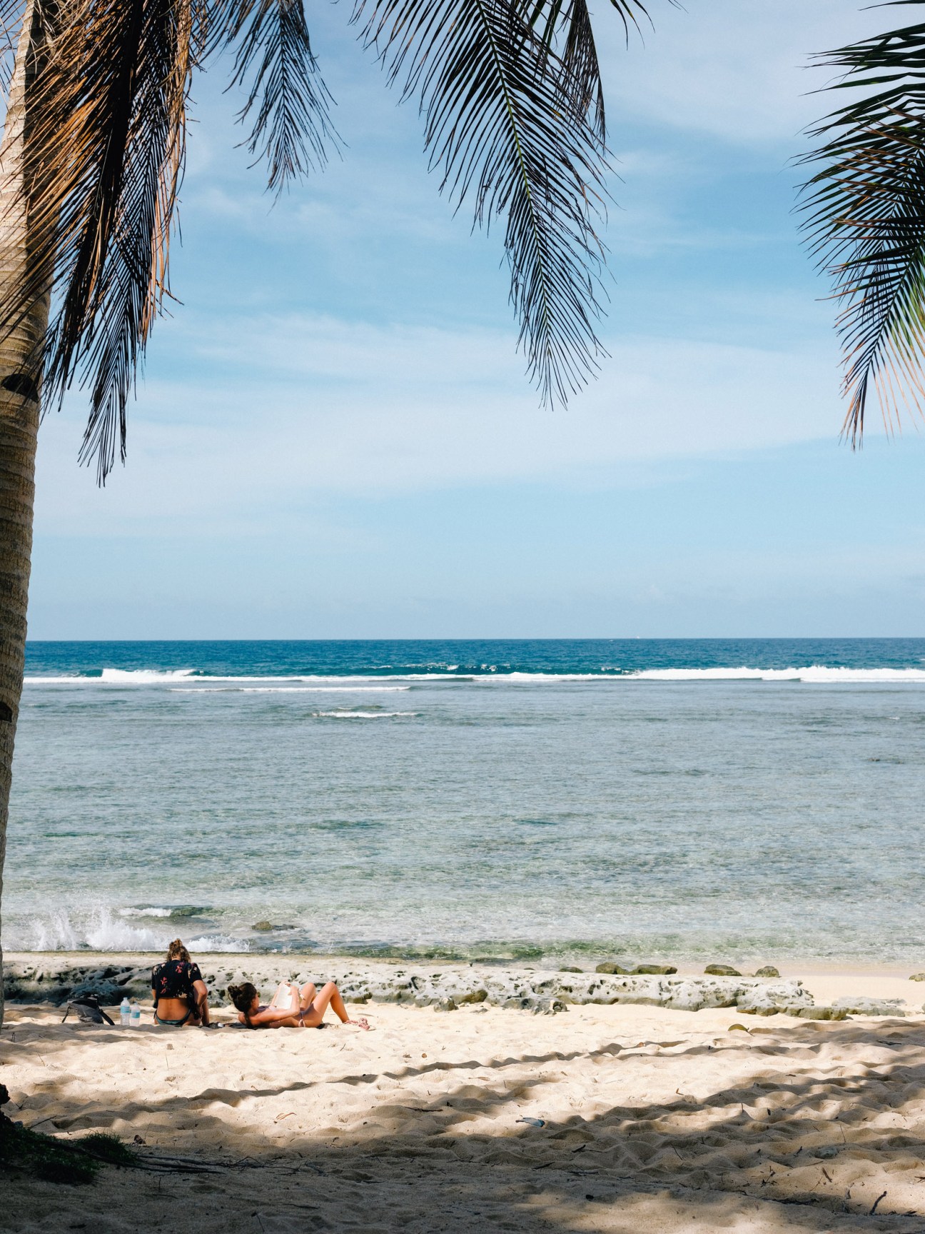 Two people relaxing on sandy beach beneath palm fronds with ocean waves and blue sky in background.