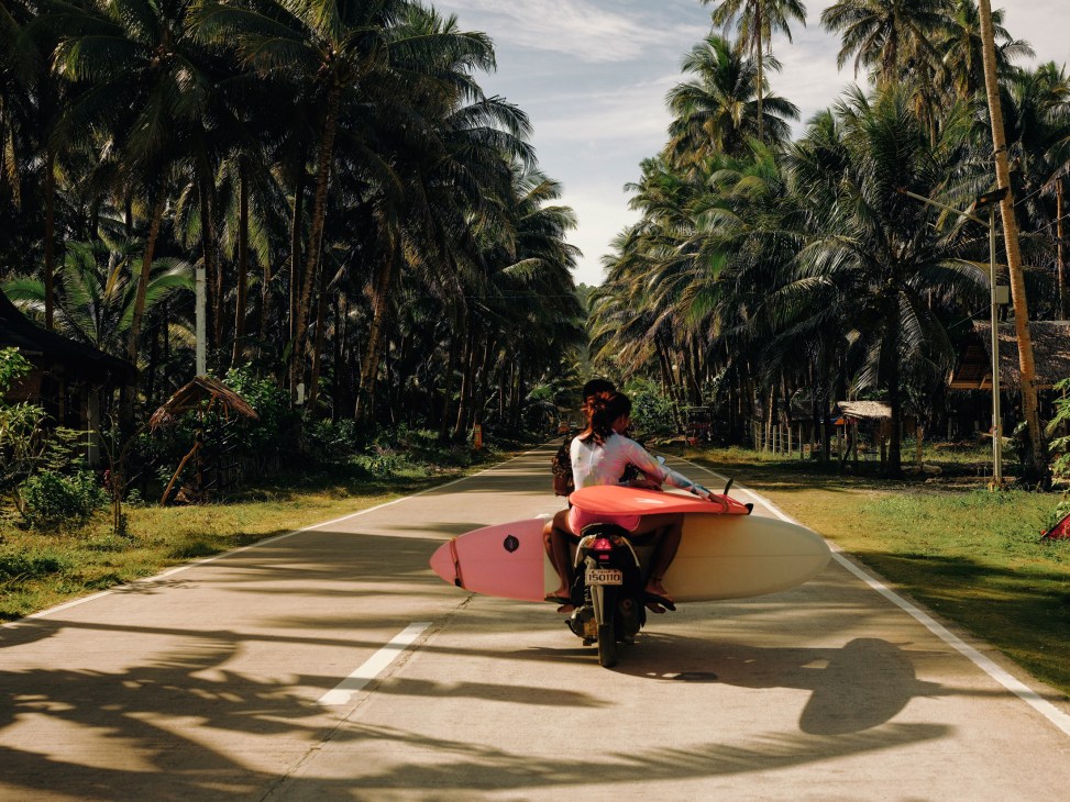 Person on a motorbike carrying a pink surfboard on a palm-lined road in Siargao, Philippines.