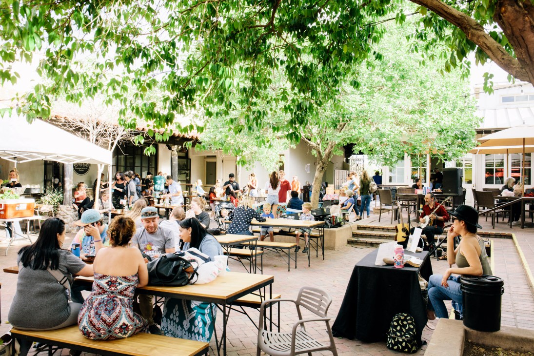 Bustling outdoor courtyard with people dining and socializing under large shade trees at the Mercado District.