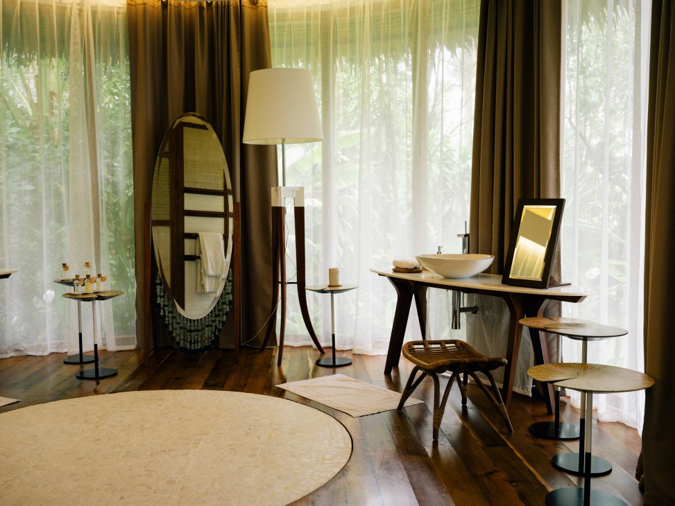 Modern resort bathroom with oval mirror, vessel sink, floor lamp, and wooden vanity overlooking tropical gardens through sheer curtains.