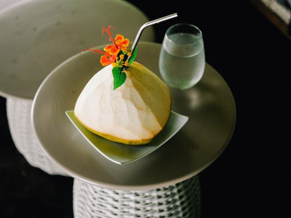 A plated dessert dome with white cream topping, yellow fruit base, and orange flowers garnish, served with a glass of green liquid on white dinnerware.